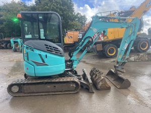 Mini excavator in turquoise color with a hydraulic arm and tracks, parked on a construction site.